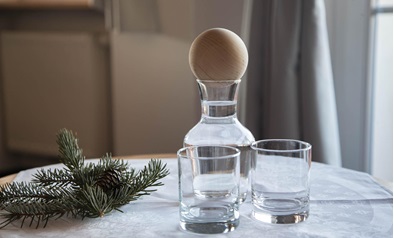 Wasserkaraffe mit Holzdeckel und zwei Gläsern auf gedecktem Tisch mit Tannenzweig.