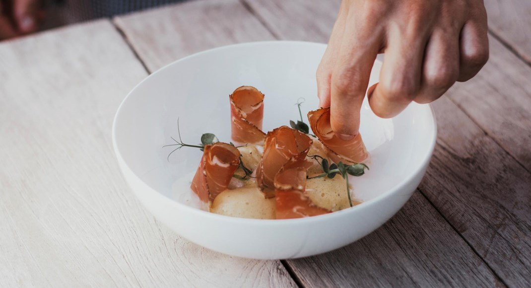 Hand arranges fine dish with slices of bacon and herbs on white plate.