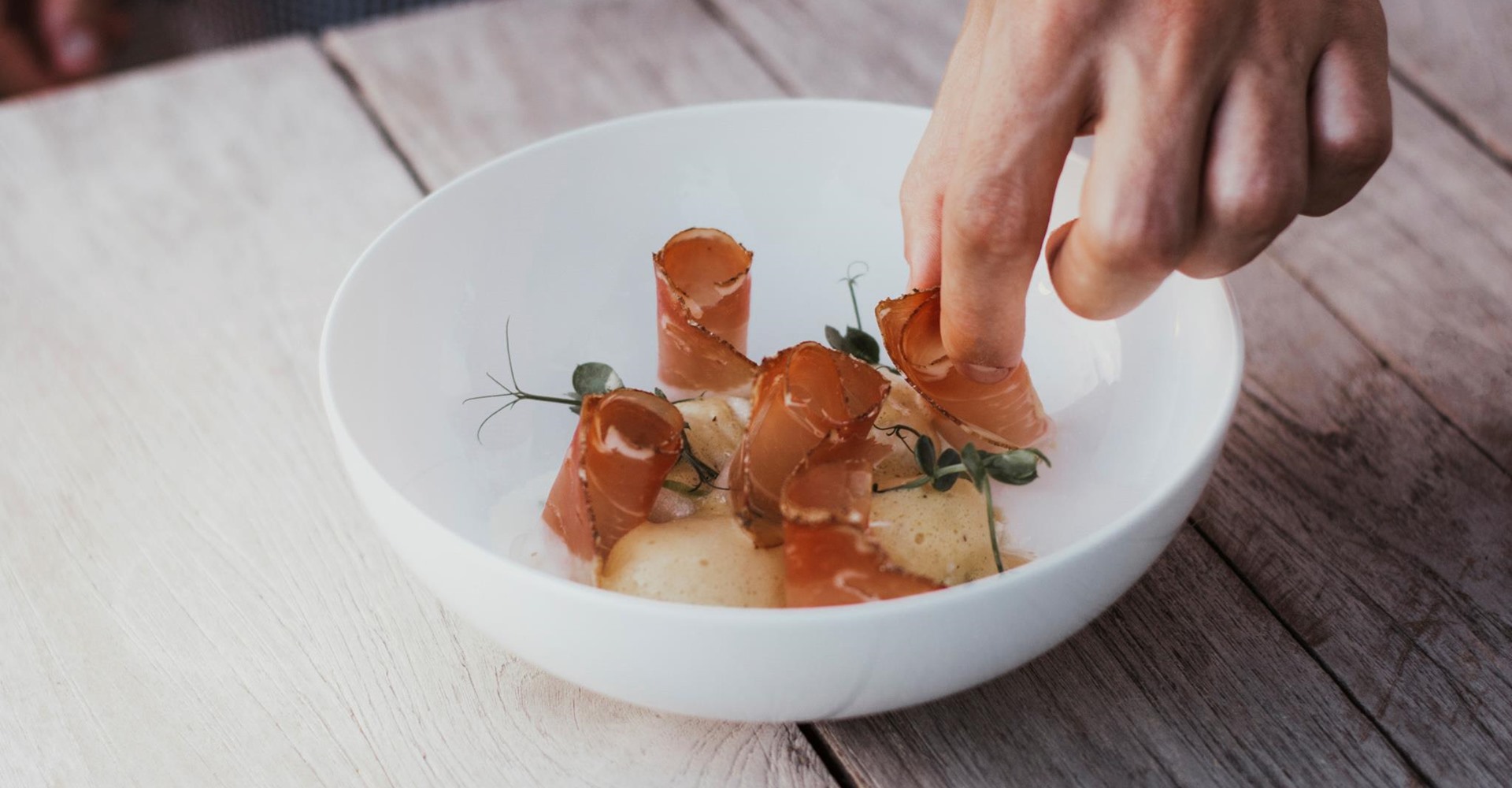 Hand arranges fine dish with slices of bacon and herbs on white plate.