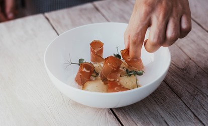 Hand arranges fine dish with slices of bacon and herbs on white plate.