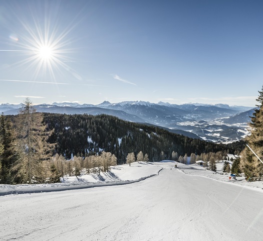 Sunny ski slope with view of forested hills and snow-covered mountains in the distance.