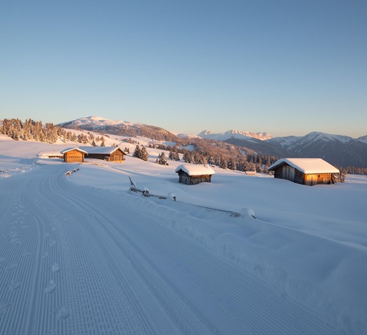 Snowy alpine huts at sunset, surrounded by mountains and freshly groomed cross-country track.