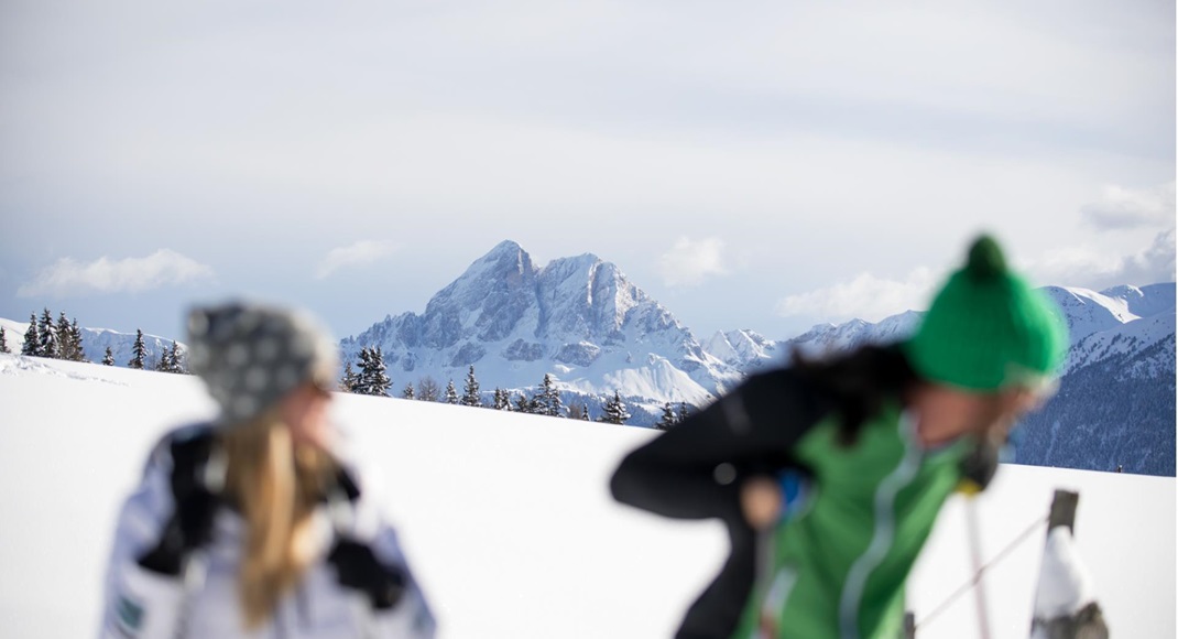 Snowy mountain landscape with a prominent peak in focus; in the foreground, two cross-country skiers out of focus in bright winter sunshine.