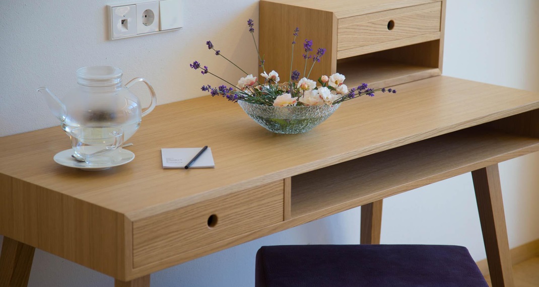 Schlichter Holztisch mit einem violetten Hocker, einer gläsernen Teekanne mit Tasse, einem Notizblock mit Stift und einer Schale mit Lavendel und kleinen weißen Blüten.