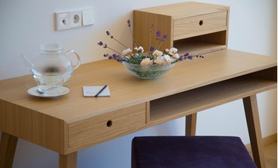 Scrivania in legno semplice con sgabello viola, teiera e tazza in vetro, blocco note con penna e ciotola con lavanda e piccoli fiori bianchi.
