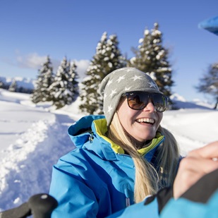 Donna sorridente con giacca invernale blu e berretto con stelle durante una passeggiata invernale, con alberi innevati sullo sfondo.