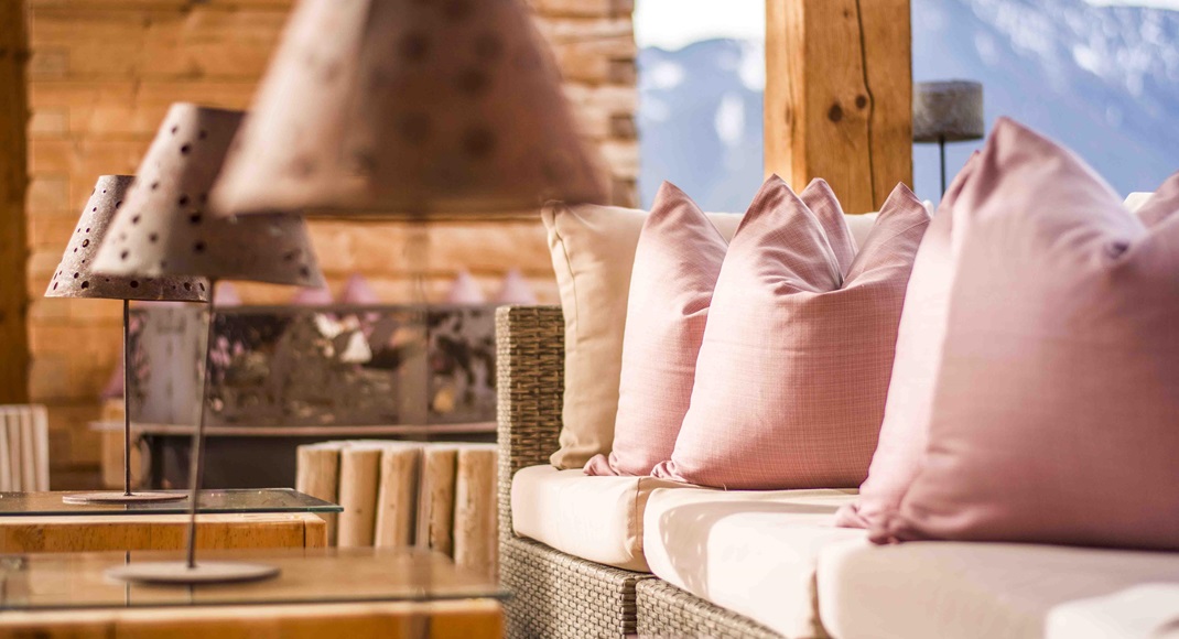 Zona lounge con cuscini rosa e vista sulle montagne, su una terrazza soleggiata in legno.