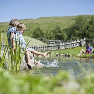 Zwei Kinder spielen am Ufer eines Naturteichs und plantschen mit den Füßen im Wasser; im Hintergrund sitzt ein Paar entspannt auf der Wiese
