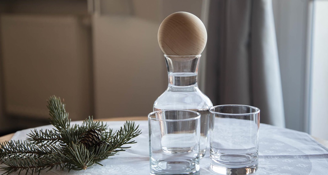 Caraffa d’acqua con tappo in legno e due bicchieri su tavolo decorato con ramo di pino.