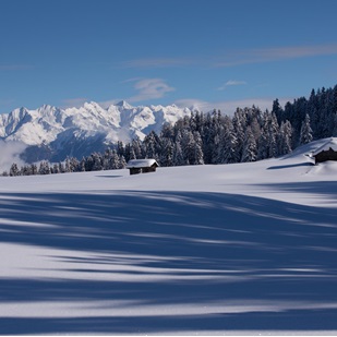 Baite innevate su una radura d'inverno con ombre sulla neve e montagne sullo sfondo.