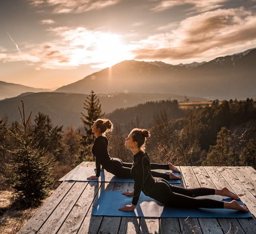 Two women doing yoga at sunrise on a wooden platform in the mountains.
