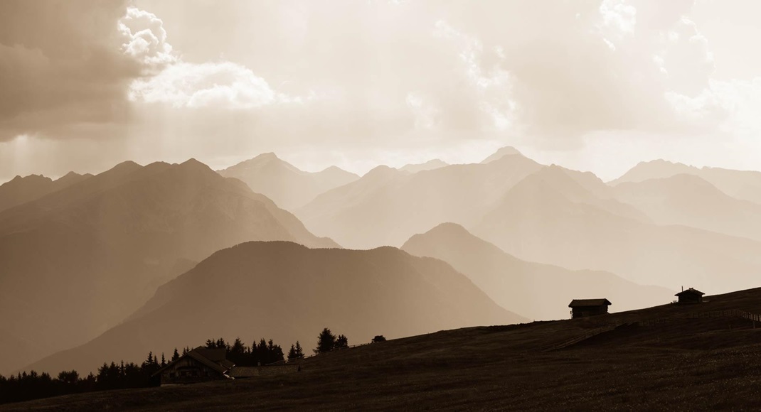 Hütten auf Almwiese vor mehreren Bergketten im Dunst und sepiafarbenes Licht.
