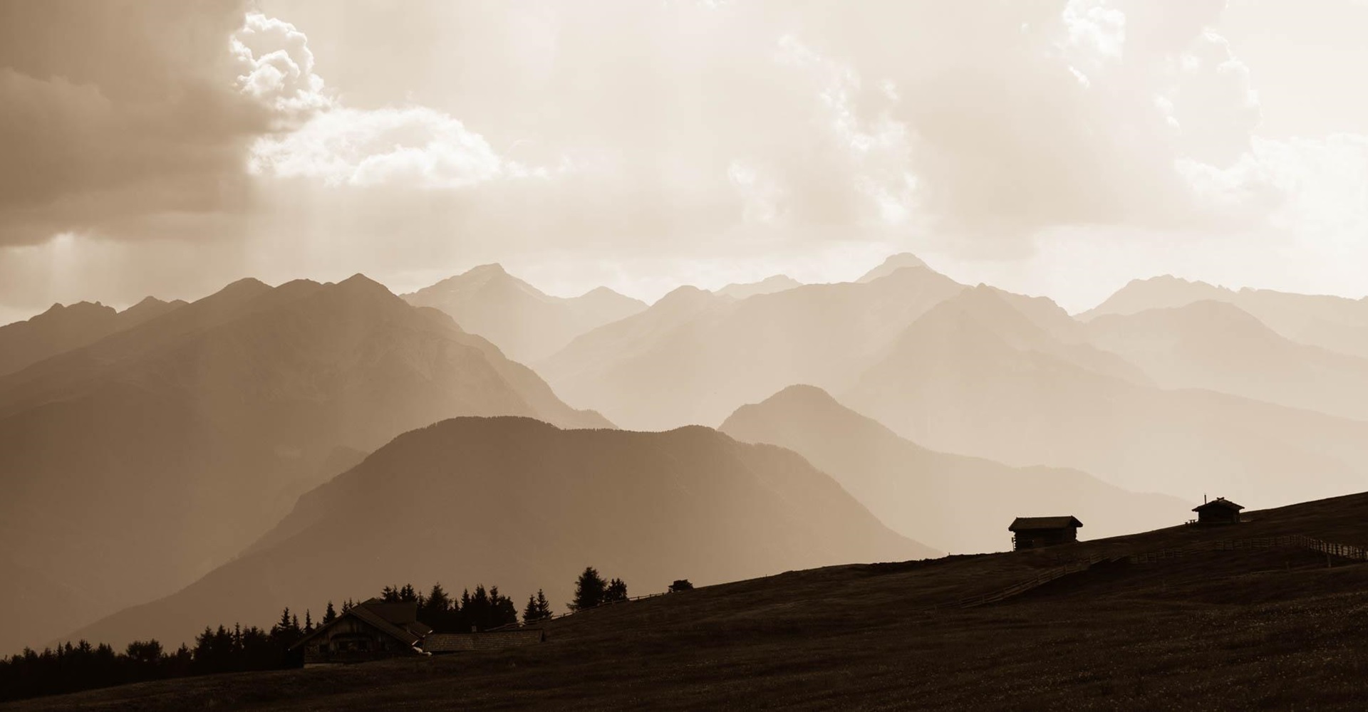 Hütten auf Almwiese vor mehreren Bergketten im Dunst und sepiafarbenes Licht.