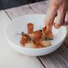Hand arranges fine dish with slices of bacon and herbs on white plate.