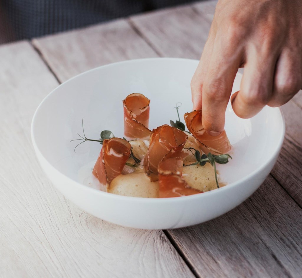 Hand arranges fine dish with slices of bacon and herbs on white plate.