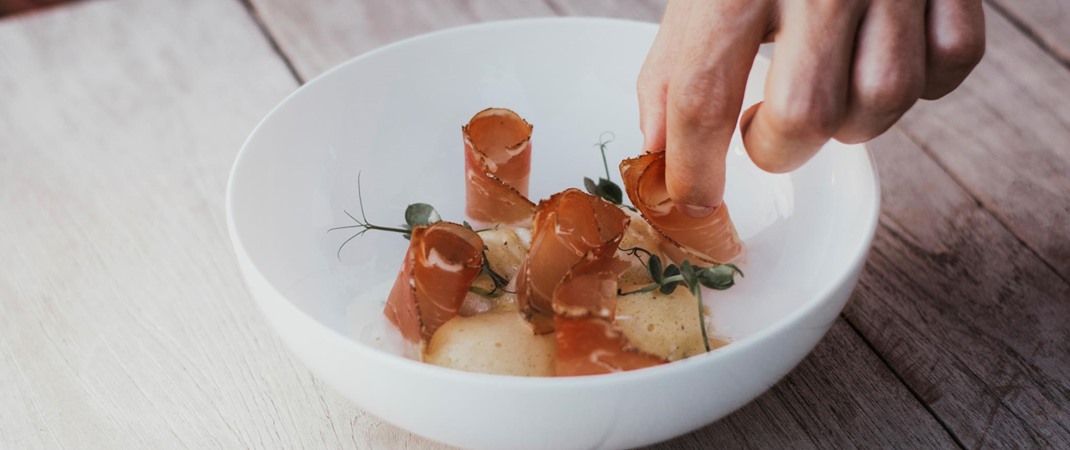 Hand arranges fine dish with slices of bacon and herbs on white plate.
