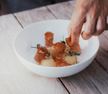 Hand arranges fine dish with slices of bacon and herbs on white plate.