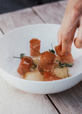 Mano impiatta un piatto raffinato con fette di speck e erbe aromatiche.