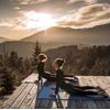 Zwei Frauen machen Yoga bei Sonnenaufgang auf einer Holzplattform in den Bergen.