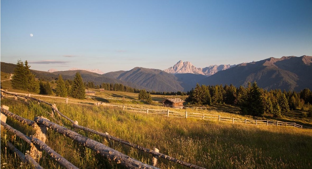 Wide alpine meadow with wooden fences and huts at sunset with mountain backdrop.