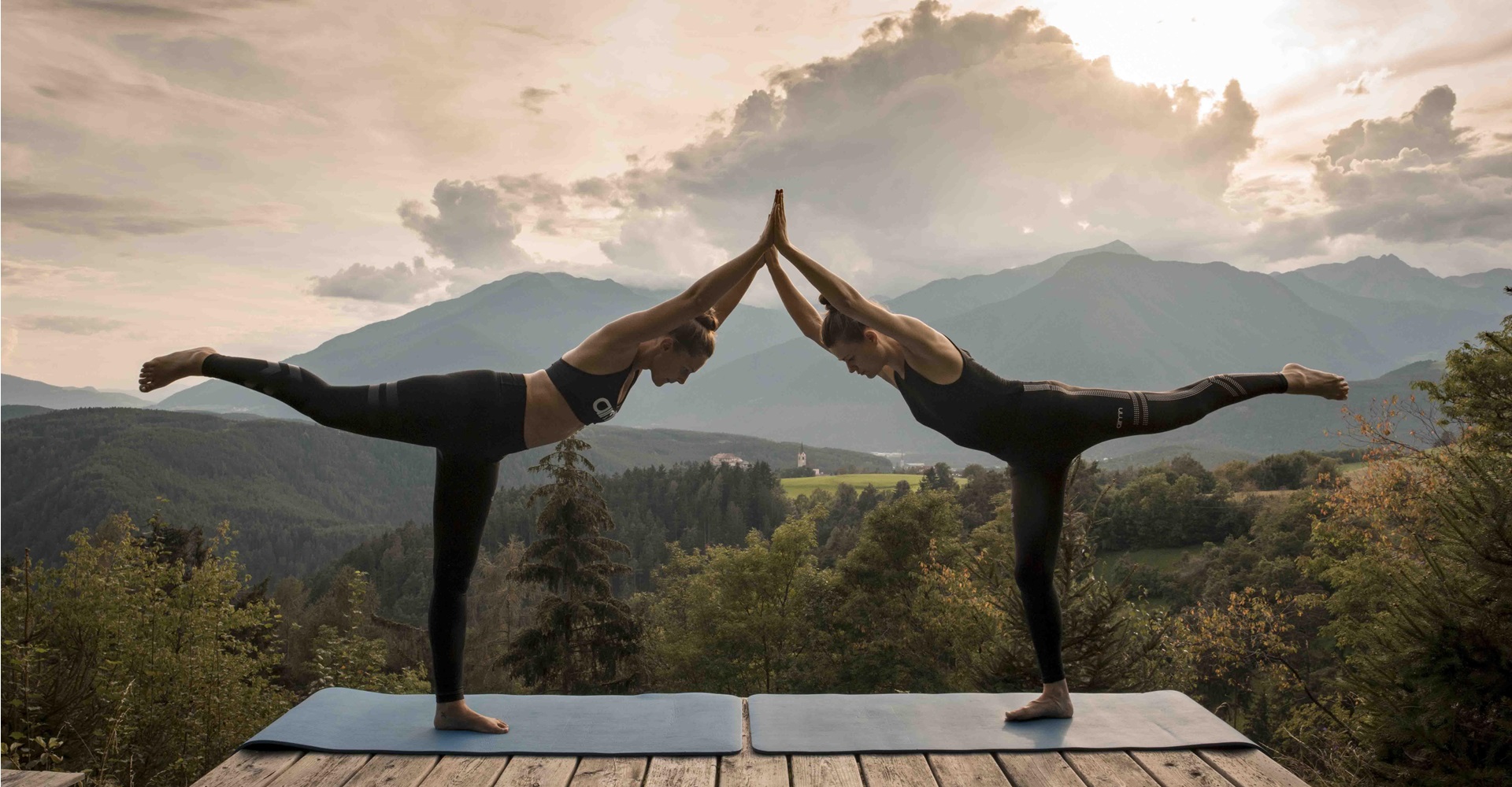 Zwei Frauen machen Yoga in Balancehaltung auf Terrasse vor Bergkulisse bei Sonnenuntergang.