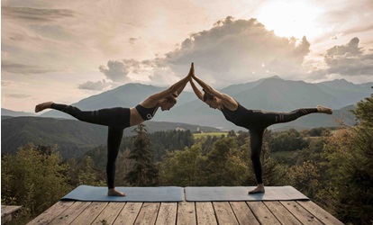Zwei Frauen machen Yoga in Balancehaltung auf Terrasse vor Bergkulisse bei Sonnenuntergang.