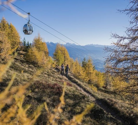 Wanderer im Herbstwald unter einer Seilbahn mit Blick auf das weite Bergpanorama.