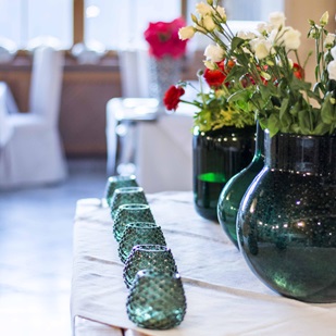 Vasen mit bunten Blumen und grüne Teelichter auf einem Holztisch, dahinter ein helles Fenster.