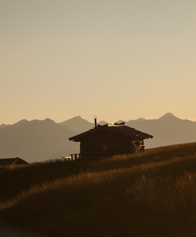 Berghütte im Gegenlicht bei Sonnenuntergang, sanfte Hügel und Silhouetten der Berge im Hintergrund.
