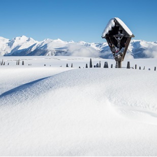 Paesaggio alpino innevato con una croce di legno in primo piano e montagne innevate sullo sfondo