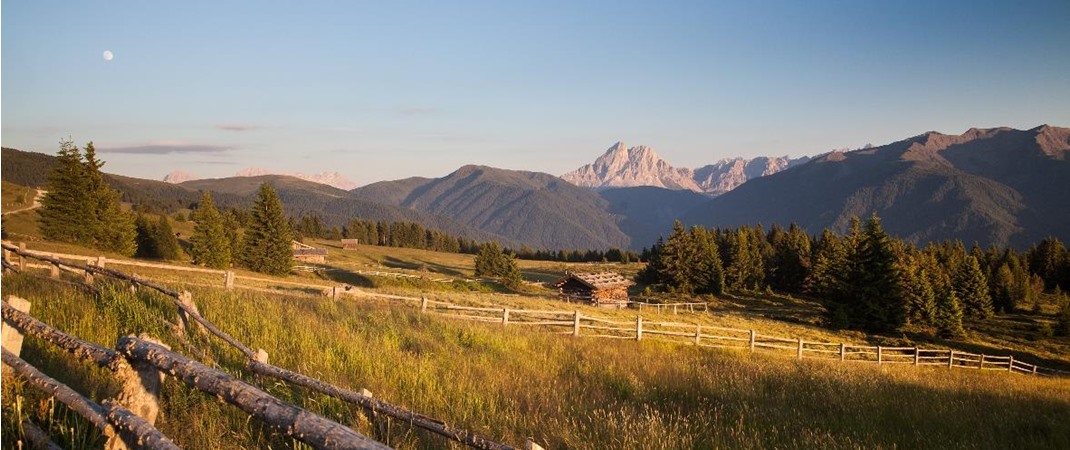 Ampio prato alpino con recinzioni in legno e baite al tramonto davanti alle montagne.