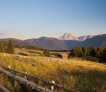 Weite Almwiese mit Holzzäunen und Hütten vor Bergkulisse bei Sonnenuntergang.