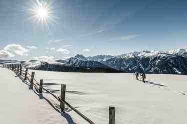 Two snowshoers in a wide winter landscape with views of snow-covered mountains under the shining sun.