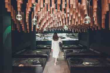 Woman in bathrobe enters a stylish relaxation room with wooden décor and loungers.