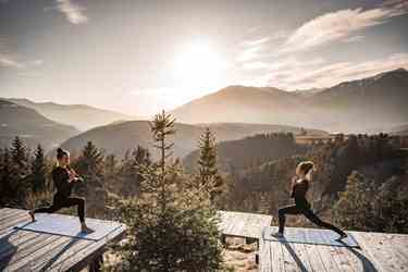 Zwei Frauen machen Yoga bei Sonnenaufgang auf einer Holzplattform in den Bergen.