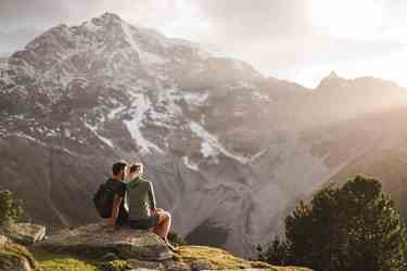 Hiking couple sits on rock, gazing at snow-dusted mountain scenery in warm sunset light.