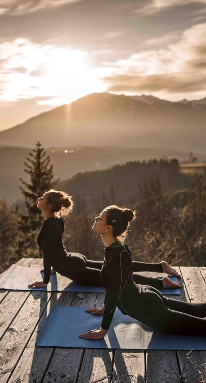 Two women doing yoga at sunrise on a wooden platform in the mountains.